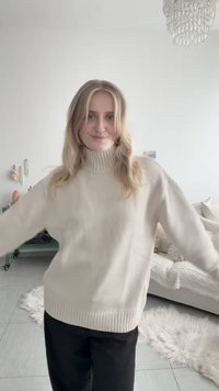 Femme aux cheveux blonds portant un pull à col roulé beige et un pantalon noir, debout dans un salon lumineux et accueillant avec un chandelier et un tapis blanc.