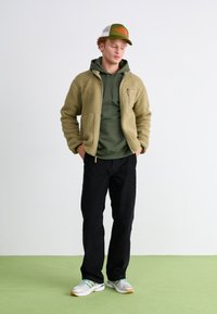Chaqueta forrada de forro polar en verde oliva, combinada con una sudadera verde oscuro, pantalones negros y zapatillas grises, rematada con una gorra verde y blanca.
