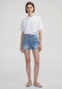 Witte kortekratige shirt met logo detail, gecombineerd met blauwe denim shorts met een gegrafeerd studs patroon, afgewerkt met beige ballerina's.