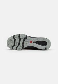 La semelle extérieure de la chaussure de sport noire présente un motif de bande de roulement profond avec du caoutchouc gris et un accent rouge. Des grips texturés améliorent la traction et la durabilité.