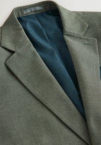 Grüner, strukturierter Blazer mit dunkelblau gemustertem Innenfutter, sichtbaren Revers und einem genähten Knopfloch im Kragenbereich.