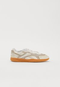 Adidași de culoare deschisă, realizati dintr-un amestec de materiale netede și texturate. Au o talpă din cauciuc de culoare portocalie, cusături vizibile și detalii cu logo.