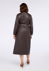 Donna che indossa un vestito midi lungo con maniche a sbuffo finto cuoio marrone scuro e sandali con tacco alto marroni e apertura sul retro, vista da dietro.