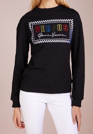 Kobieta ubrana w czarną bluzę z długim rękawem z kolorowym logo "VERSUS Gianni Versace" otoczonym szachownicowym obramowaniem, w połączeniu z białymi spodniami.