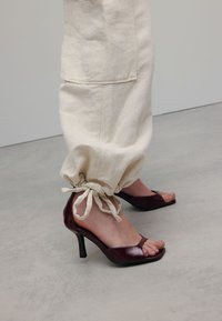 Lichte crèmekleurige joggingbroek van stof met elastische manchetten en zijzakken, gecombineerd met donker bordeauxkleurige lakleren hoge hakken met een open teen ontwerp.