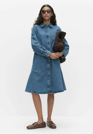 Kobieta ubrana w długą sukienkę z denimu z długim rękawem, brązowe mokasyny, okulary przeciwsłoneczne i trzymająca brązową, fakturowaną kopertówkę na jednolitym białym tle.