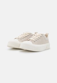 Beige lerret sneakers med kremfargede gummisåler, runde tår, og flate snører. Har en teksturert overdel og en løkke bak hælen.