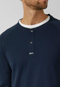 Marineblaues, geripptes Langarmshirt mit Rundhalsausschnitt, das zwei schwarze Knöpfe und einen weißen Innenkragen aufweist. Kleines Etikett an der Seite.
