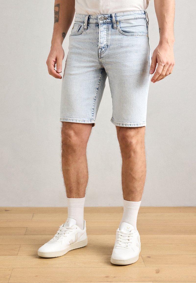 Shorts en denim bleu clair avec cinq poches, une taille mi-haute et des ourlets roulés, associés à des baskets blanches et des chaussettes de longueur mi-mollet.