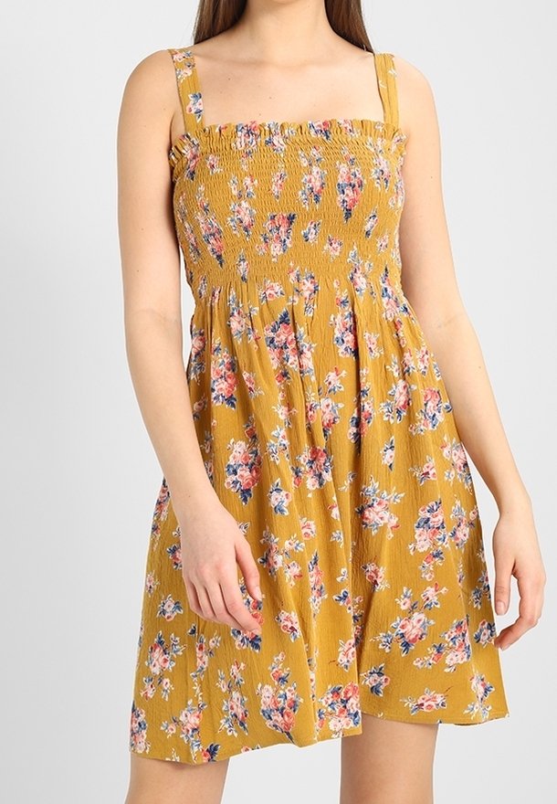 Femme portant une robe d'été sans manches couleur jaune moutarde avec un imprimé floral rose et bleu, un corsage smocké et un décolleté carré.