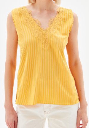 Kvinde iført gulstripet ærmeløs top med blonden V-udskæring og hvide bukser, stående mod en hvid baggrund.