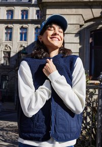 Jeune femme aux cheveux foncés portant une casquette bleue, un gilet bleu marine et une chemise blanche à manches longues souriante en extérieur devant un bâtiment historique.