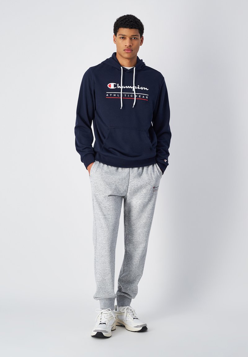 Felpa della marina con cordini bianchi e logo; pantaloni della tuta grigi con polsini; scarpe da ginnastica bianche. Abbigliamento sportivo semplice e casual.