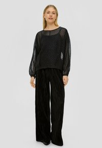 Schwarze transparente Bluse mit Tupfen, ausgestattet mit langen Puffärmeln, kombiniert mit weit geschnittenen, gefalteten schwarzen Hosen, die eine lockere Silhouette schaffen.