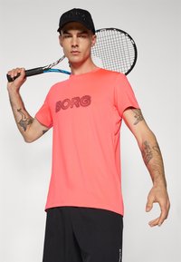 Korallfarget treningstrøye med "BORG" i fet svart tekst, kombinert med svarte shorts og en matchende caps, holdt sammen med en tennisracket.