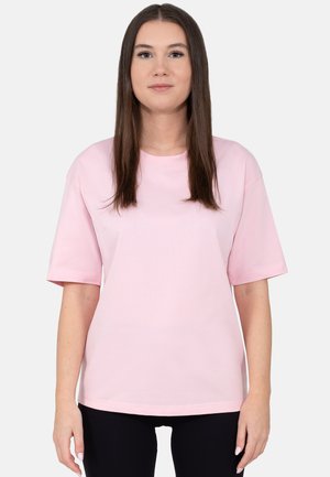 Young woman with long brown hair wearing a light pink short-sleeve t-shirt and black pants, standing against a plain white background.