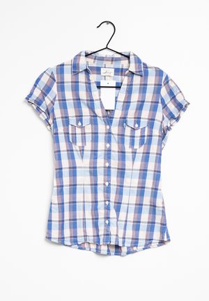 Camisa de manga corta a cuadros en azul y blanco con botones y dos bolsillos en el pecho, colgada en una percha negra contra un fondo blanco.