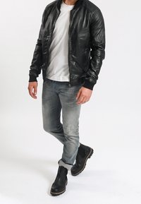 Man som bär svart läderjacka, vit t-shirt, urtvättade jeans och svarta stövlar, går mot en helt vit bakgrund.