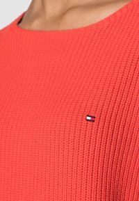 Gros plan sur un vêtement en tricot texturé rouge avec un petit logo rectangulaire rouge, blanc et bleu sur la zone de la poitrine.