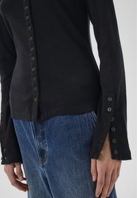 Čierny rebrovaný top s dlhým rukávom a gombíkmi na prednej strane a na manžetách, nosený so modrými rifľami.