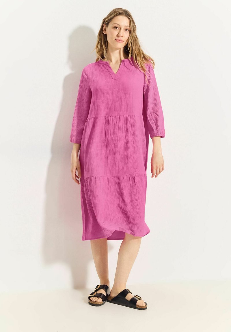 Rosa, knielanges Kleid mit V-Ausschnitt, Dreiviertelärmel und gestuftem Design aus strukturiertem Stoff. Getragen mit schwarzen Sandalen.
