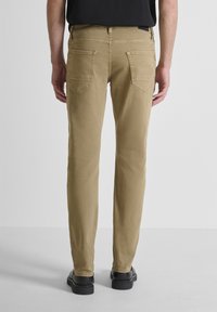 Hombre de pie con pantalones ajustados color beige y zapatos negros, visto desde atrás contra un fondo claro liso.