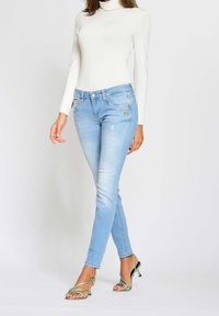 Jeans de mezclilla azul claro con corte slim, que presentan desgastes sutiles, efecto de desgaste y bolsillos frontales. Combinados con una blusa blanca ajustada y tacones verdes.