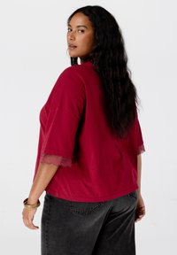 Vrouw met lange donkere haren in een bordeauxrode top met kantafgezette mouwen en zwarte jeans, die over haar schouder kijkt tegen een witte achtergrond.