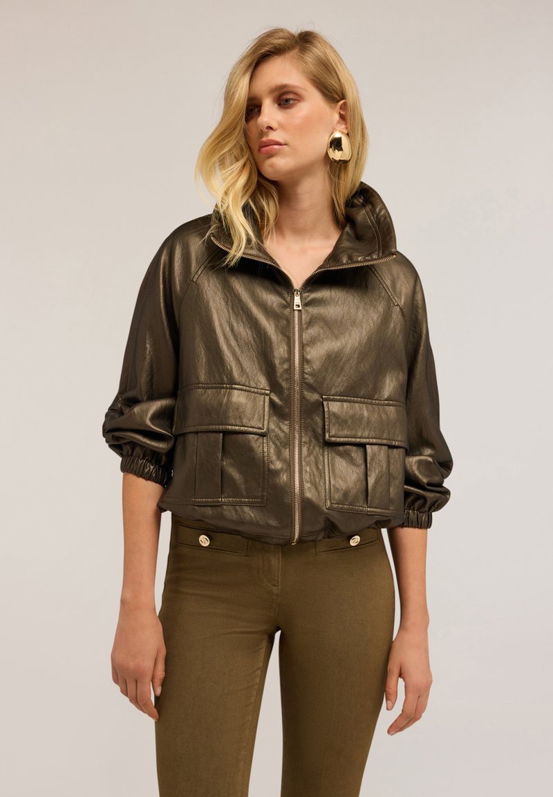 Giacca imbottita in bronzo metallico con colletto alto, chiusura frontale con zip e due grandi tasche anteriori. Presenta polsini elasticizzati e una vestibilità comoda.