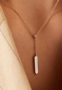 Le collier en chaîne argentée présente un pendentif géométrique avec une surface polie et blanche, accentuée par un petit détail transparent au-dessus du pendentif.