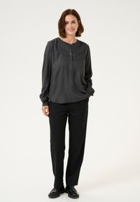 Donkergrijze blouse met lange mouwen, een ronde halslijn, knopen aan de voorkant en geplooide schouders, gecombineerd met zwarte broek en zwarte schoenen.