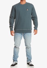 Felpa grigia con scollo rotondo e una piccola etichetta logo sul petto, abbinata a jeans blu chiaro e scarpe nere.