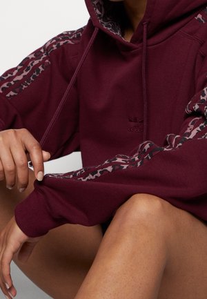 Personne portant un sweat à capuche Adidas bordeaux avec des rayures à motifs sur les manches, assise les mains reposant sur les genoux.