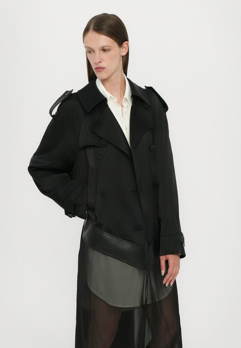 Cappotto lungo nero con un design strutturato, caratterizzato da bottoni doppi, spalline e un strato inferiore trasparente.