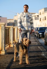 Weiß und grau gestreifte Fleecejacke, graue Hose, Mann, der einen braunen und schwarzen Hund auf Kopfsteinpflaster führt, städtische Umgebung, klarer Himmel.