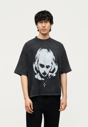 Νέος με μαύρα σγουρά μαλλιά, που φοράει φαρδιά μαύρη μπλούζα με λευκό γραφικό ενός προσώπου που μοιάζει με κρανίο με σκοτεινές κόγχες ματιών και κολιέ.