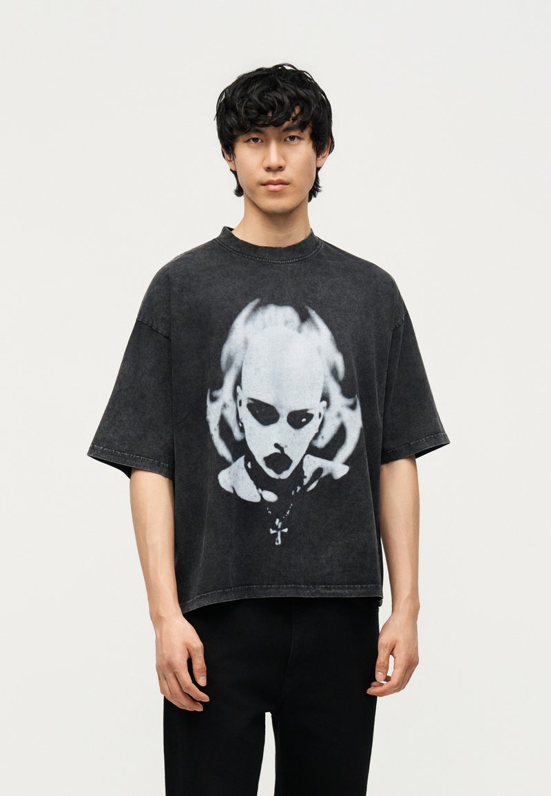 Νέος με μαύρα σγουρά μαλλιά, που φοράει φαρδιά μαύρη μπλούζα με λευκό γραφικό ενός προσώπου που μοιάζει με κρανίο με σκοτεινές κόγχες ματιών και κολιέ.