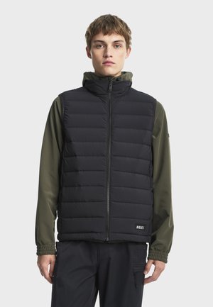 Personne portant un gilet matelassé noir par-dessus une veste à capuche vert olive, debout devant un fond uni gris clair.