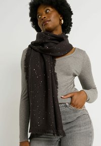 Écharpe noire avec des sequins argentés, faite de tissu doux, avec des bords effilochés. Portée de manière décontractée sur un haut à manches longues gris.