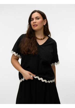 Mujer con cabello largo y castaño, vestida con blusa negra de escote en V y falda con borde ondulado blanco, sosteniendo el dobladillo, sobre un fondo claro y sencillo.