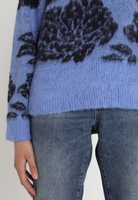 Modrý pletený svetr s černým květinovým vzorem, s ribbovanými manžetami a lemem, nošený přes světle modré džíny. Měkká textura, volný střih.