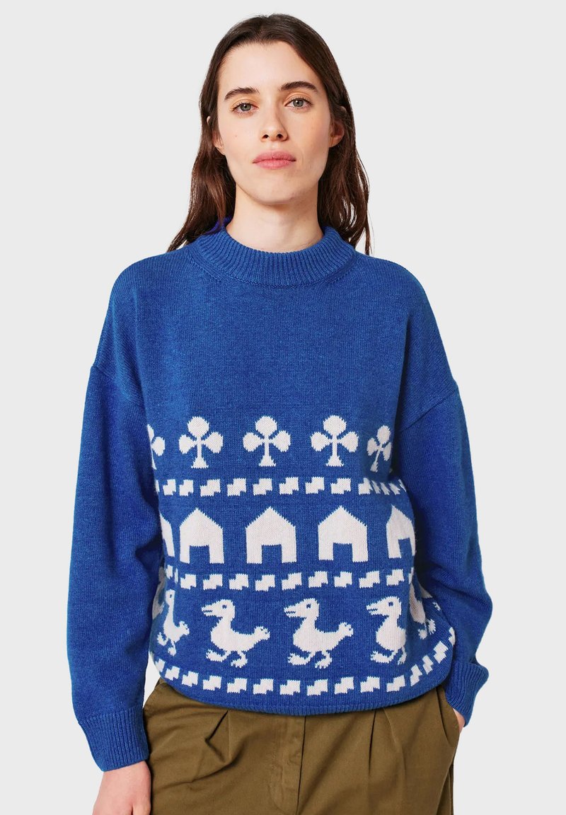 Frau, die einen blauen Pullover mit weißen Mustern von Kleeblättern, Hausformen und Vögeln trägt, kombiniert mit braunen Hosen und vor einem einfarbigen Hintergrund steht.