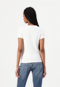Top attillato bianco a maniche corte con texture a costine, indossato con jeans blu con dettagli delle tasche posteriori. Visuale posteriore mostrata.