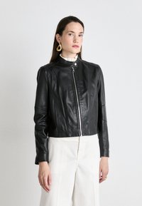 Svart läderkort jacka med hög krage, frontdragkedja och subtil sömdetaljering. Bärs över en vit blus och matchas med vida byxor.