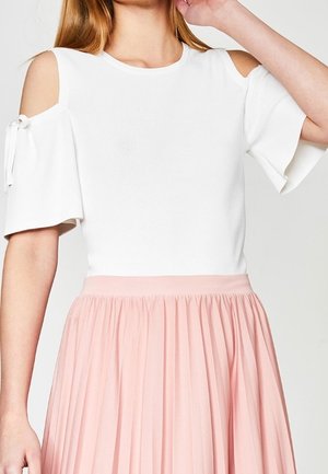 Femme portant un top blanc à épaules dénudées avec des manches courtes évasées et une jupe plissée rose clair, main près de son cou.
