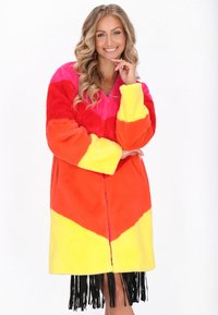 Manteau en fausse fourrure avec un motif en chevrons dans des couleurs vives : rose, orange et jaune. Il a des manches longues et un ourlet à franges.