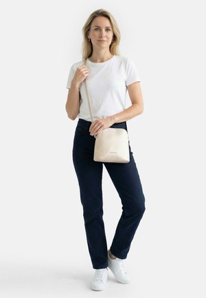 Frau steht mit beiger Umhängetasche, trägt weißes T-Shirt, dunkle Jeans, weiße Turnschuhe, neutraler Gesichtsausdruck vor hellem Hintergrund.