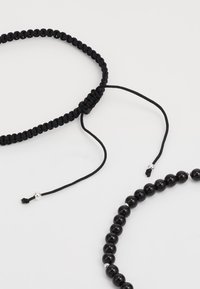 Bracciale regolabile in macramè nero con chiusura a nodo e bracciale liscio con perle lucide nere e accenti argentati.