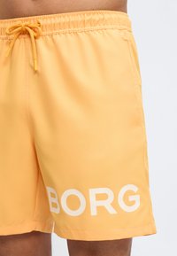 Shorts de bain orange vif avec taille élastique et cordon de serrage, arborant un grand texte blanc "BORG" sur la jambe gauche.