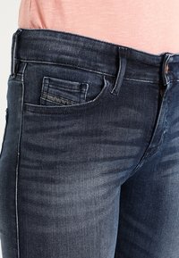 Jeans de mezclilla azul oscuro con corte ajustado, que cuentan con un pequeño bolsillo frontal y un ligero desgastado a lo largo de los muslos y la cintura.